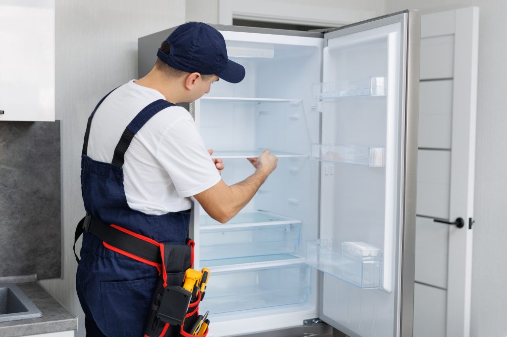 Man in a uniform with tools is repairing a refrigerator in the kitchen.