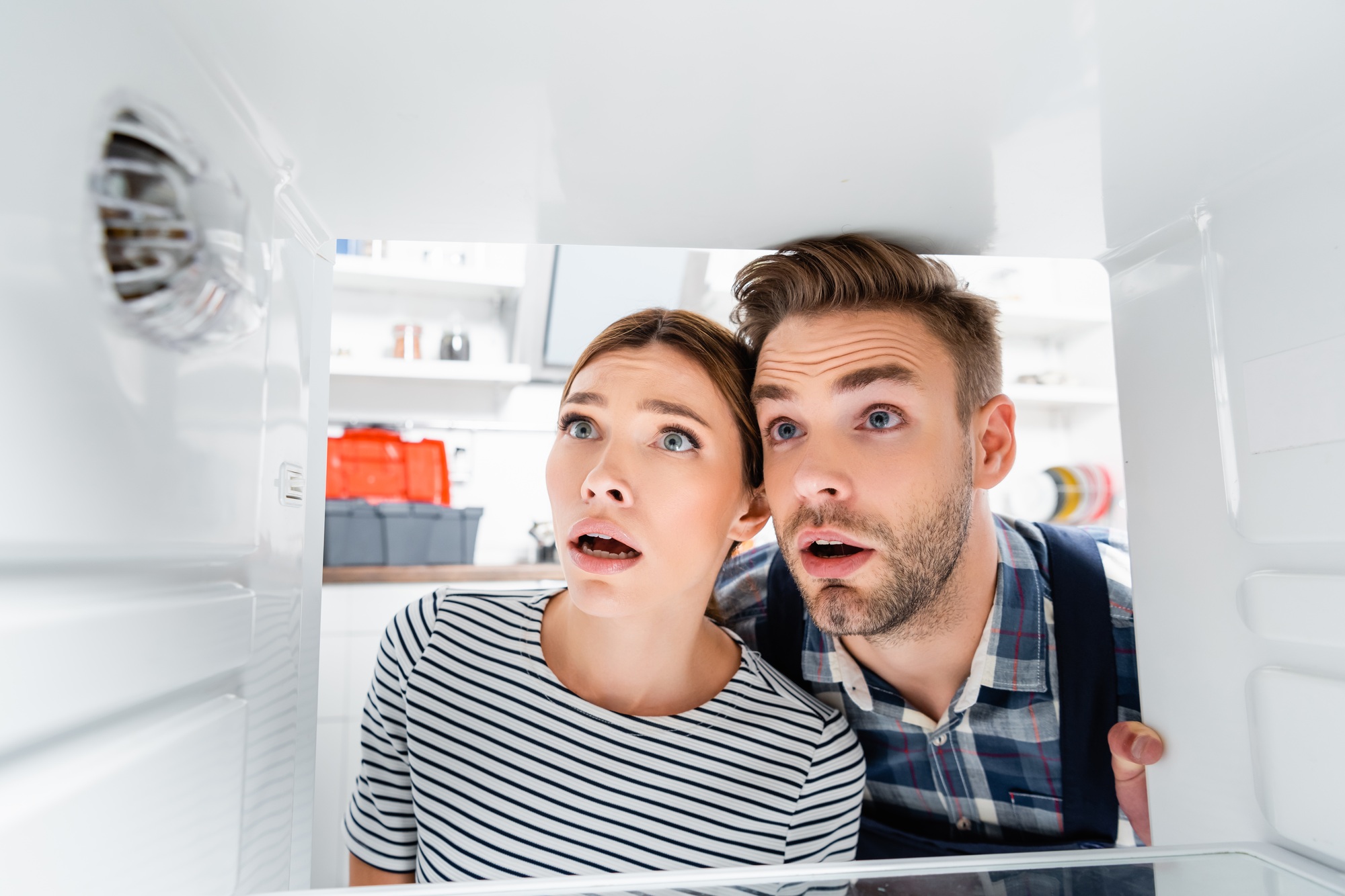 shocked young woman and handyman with open mouths looking at freezer on blurred foreground in