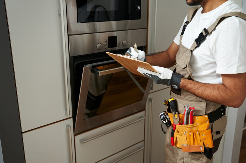 Close up of service worker in uniform standing near oven and writing on clipboard in kitchen