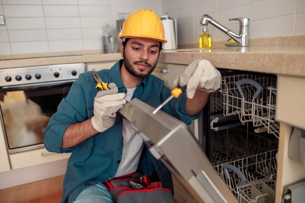 Focused handyman in special clothing repairing dishwasher in modern kitchen