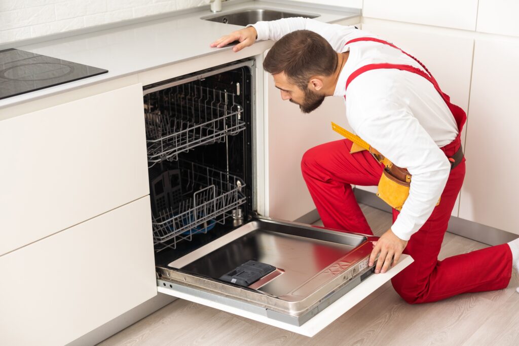 Young modern serviceman in worker suit during the repairing of the dishwasher on the domestic