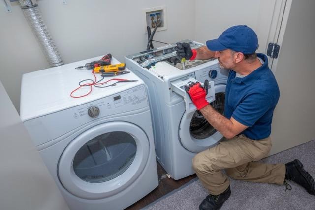 A technician wearing gloves and a blue cap kneels while repairing the top of an open washing machine next to a dryer.
