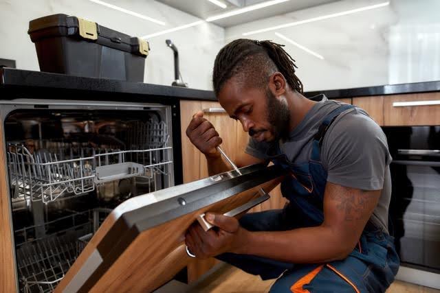 A technician kneeling in a kitchen while using a screwdriver to repair an open dishwasher.