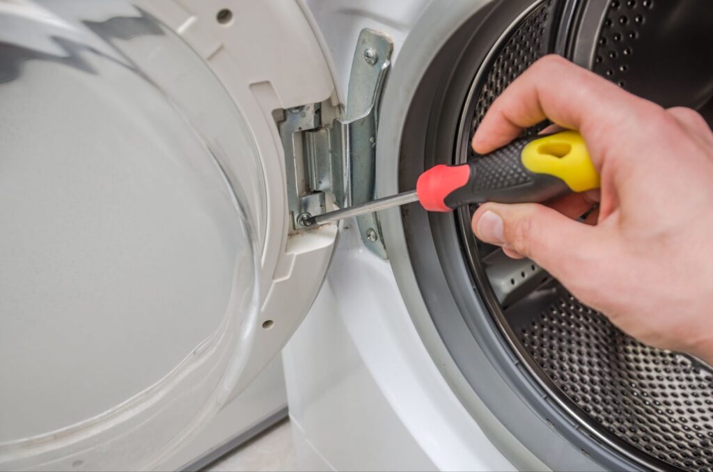 A close-up view of a hand using a screwdriver to tighten the door hinge of a white front-loading washing machine.