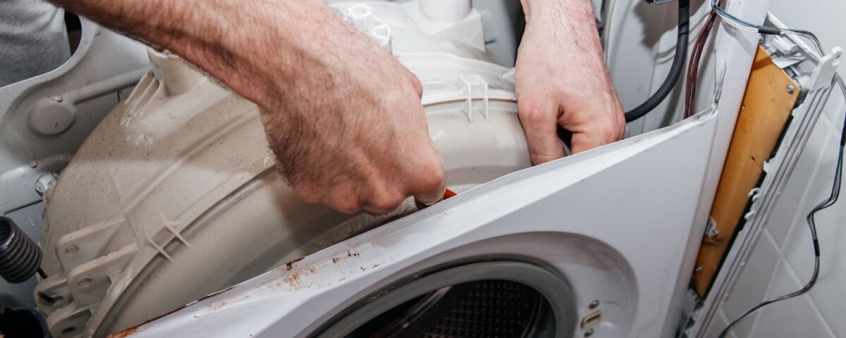 A person's hairy hands reach into the open top of a disassembled white washing machine to adjust or repair the internal drum.