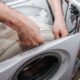 A person's hairy hands reach into the open top of a disassembled white washing machine to adjust or repair the internal drum.