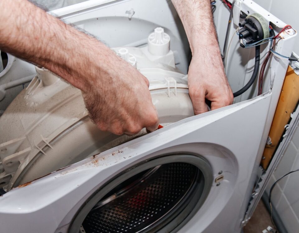 A person's hairy hands reach into the open top of a disassembled white washing machine to adjust or repair the internal drum.