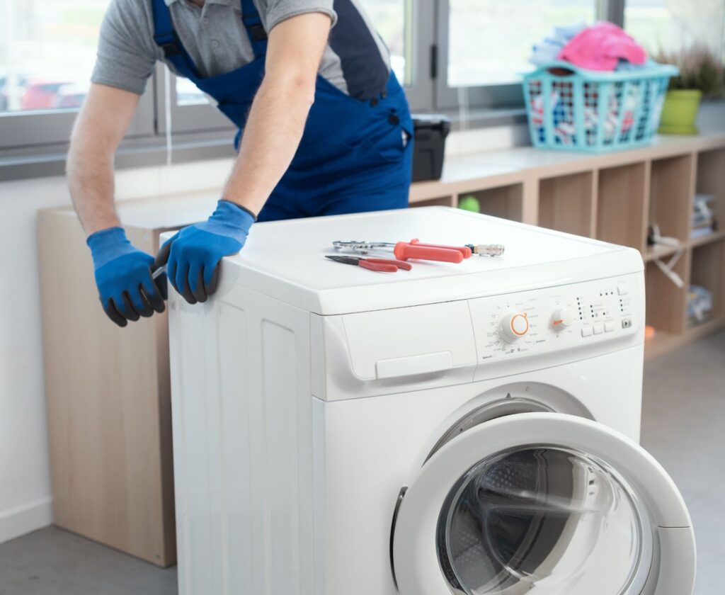 A technician in blue overalls and gloves uses tools to repair a white front-loading washing machine in a laundry room.