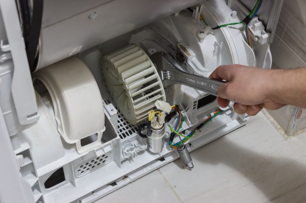 A close-up of a technicians hand using an adjustable wrench to repair or tighten a white plastic blower wheel inside the bottom compartment of a dryer.