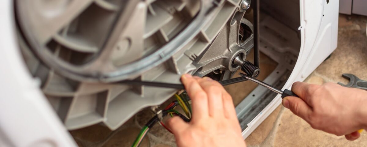 A close-up of a technician's hands using a screwdriver to repair or replace the drive belt on the back of a washing machine.