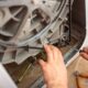 A close-up of a technician's hands using a screwdriver to repair or replace the drive belt on the back of a washing machine.
