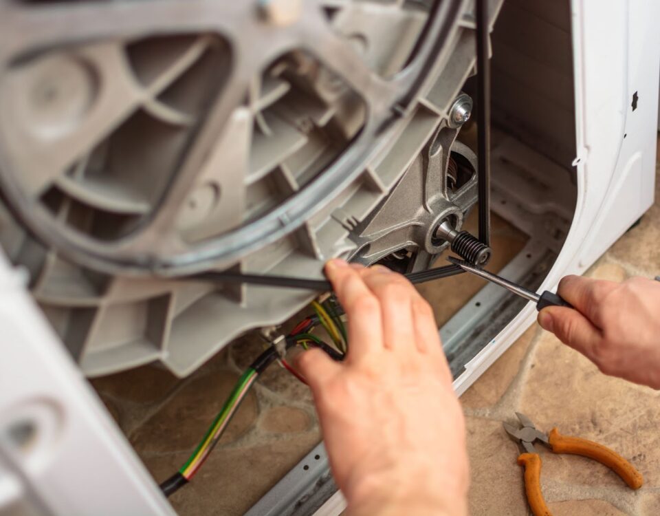 A close-up of a technician's hands using a screwdriver to repair or replace the drive belt on the back of a washing machine.