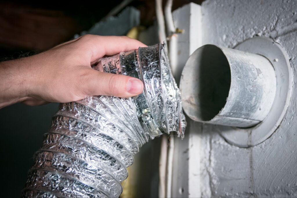 A persons hand connecting a flexible foil dryer vent hose to a metal wall exhaust pipe.