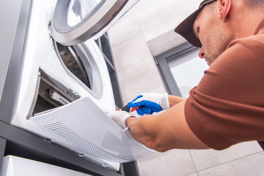 A technician in gloves using a tool to repair or maintain the bottom service panel of a front-loading washing machine.