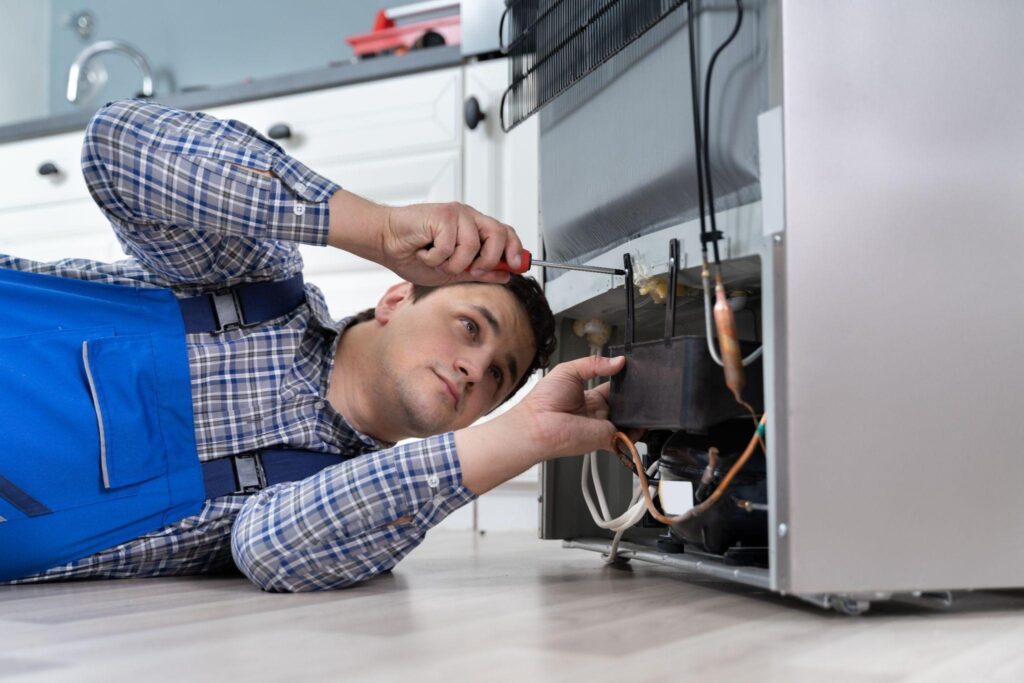 A man is positioned on the floor with his hands placed on the sides of a refrigerator