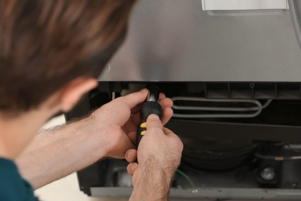 A man repairs a refrigerator using a screwdriver focused on the appliance interior components